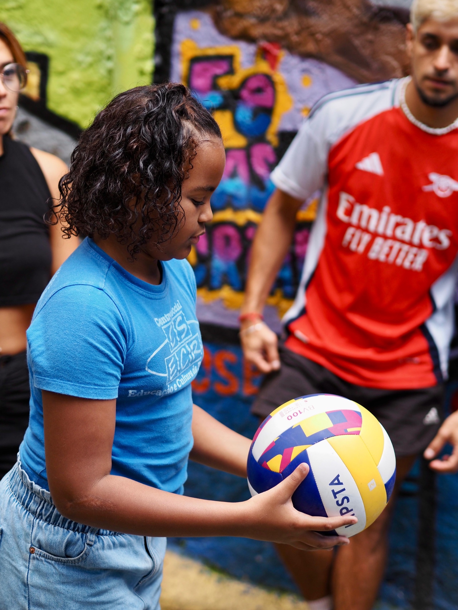 A volunteer in a football shirt passes a volleyball to a girl in front of a painted mural.