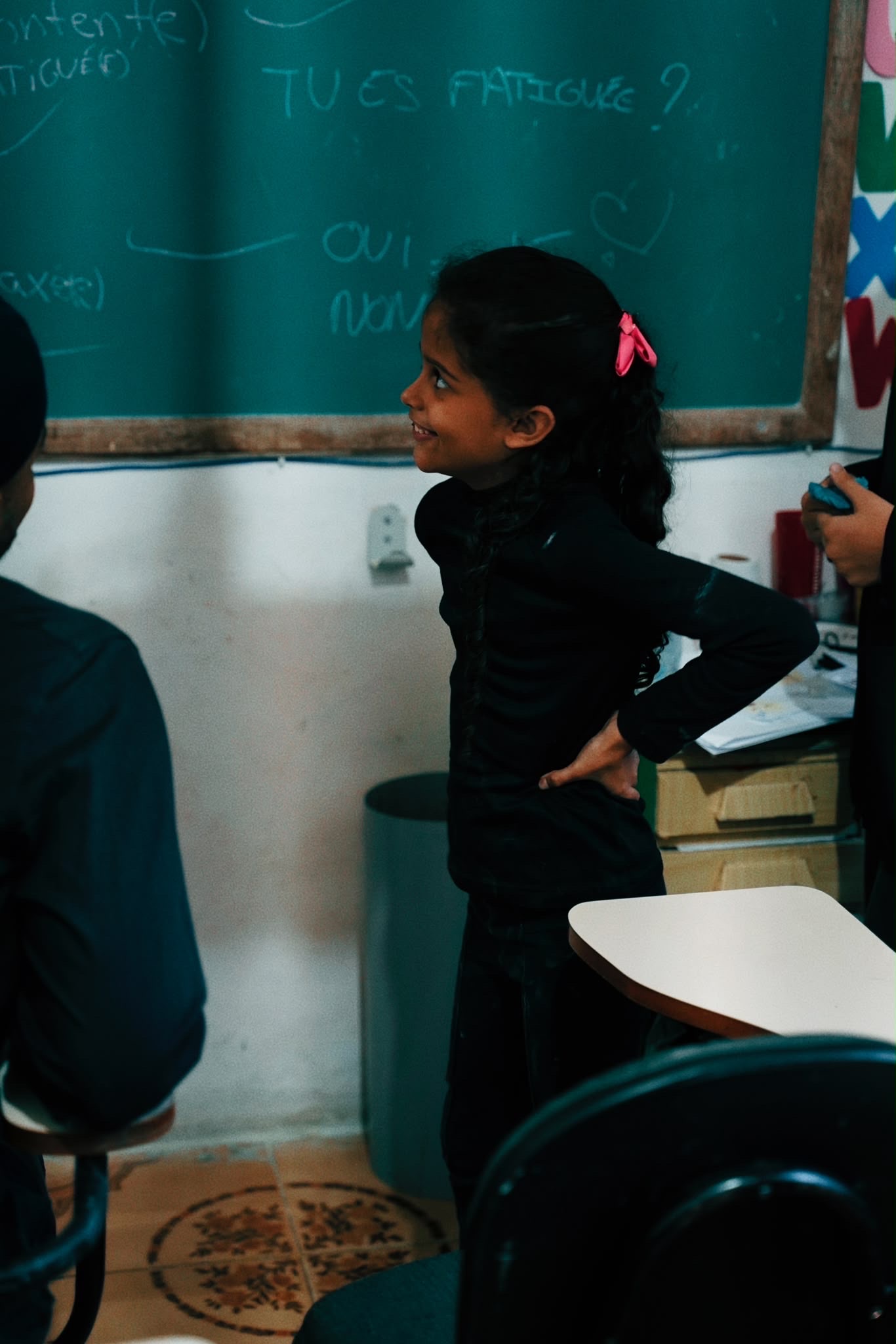 A smiling girl stands at the front of a classroom beside a chalkboard.