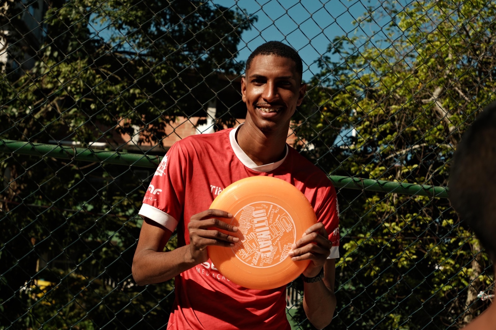 A young man in a red sports shirt smiles while holding an orange frisbee in front of a chain-link fence.