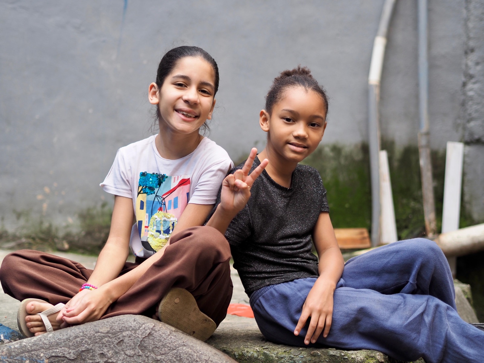 Two girls sit together on a ledge, one making a peace sign.