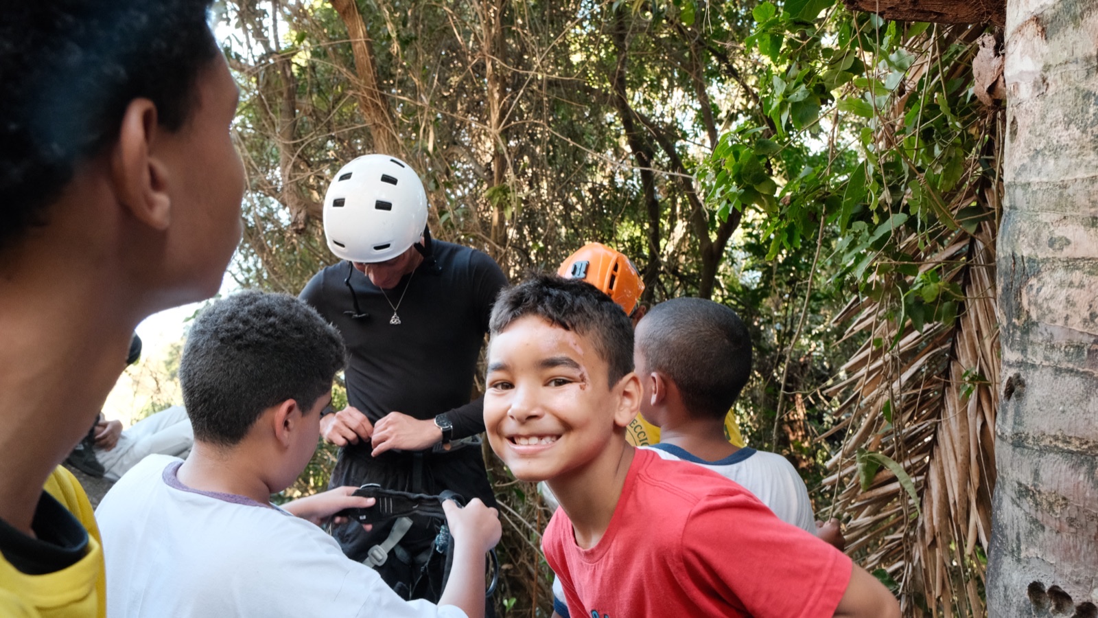 Smiling children in climbing helmets gather around an instructor on a forest trail.