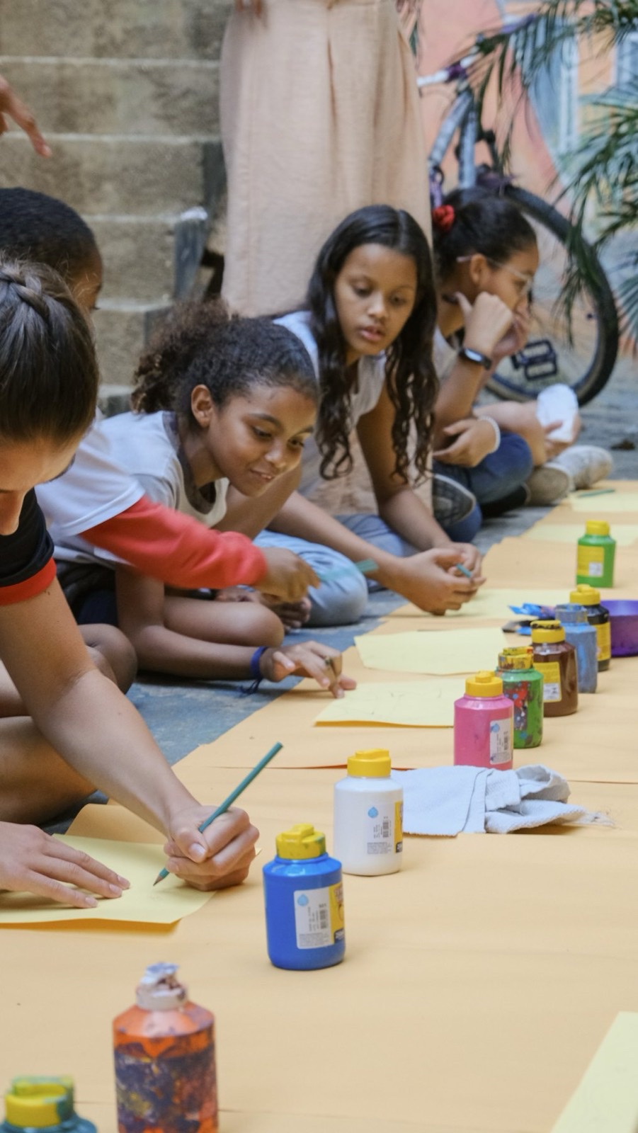 Girls kneeling along a strip of paper, painting, with bottles of colour lined up in front of them.