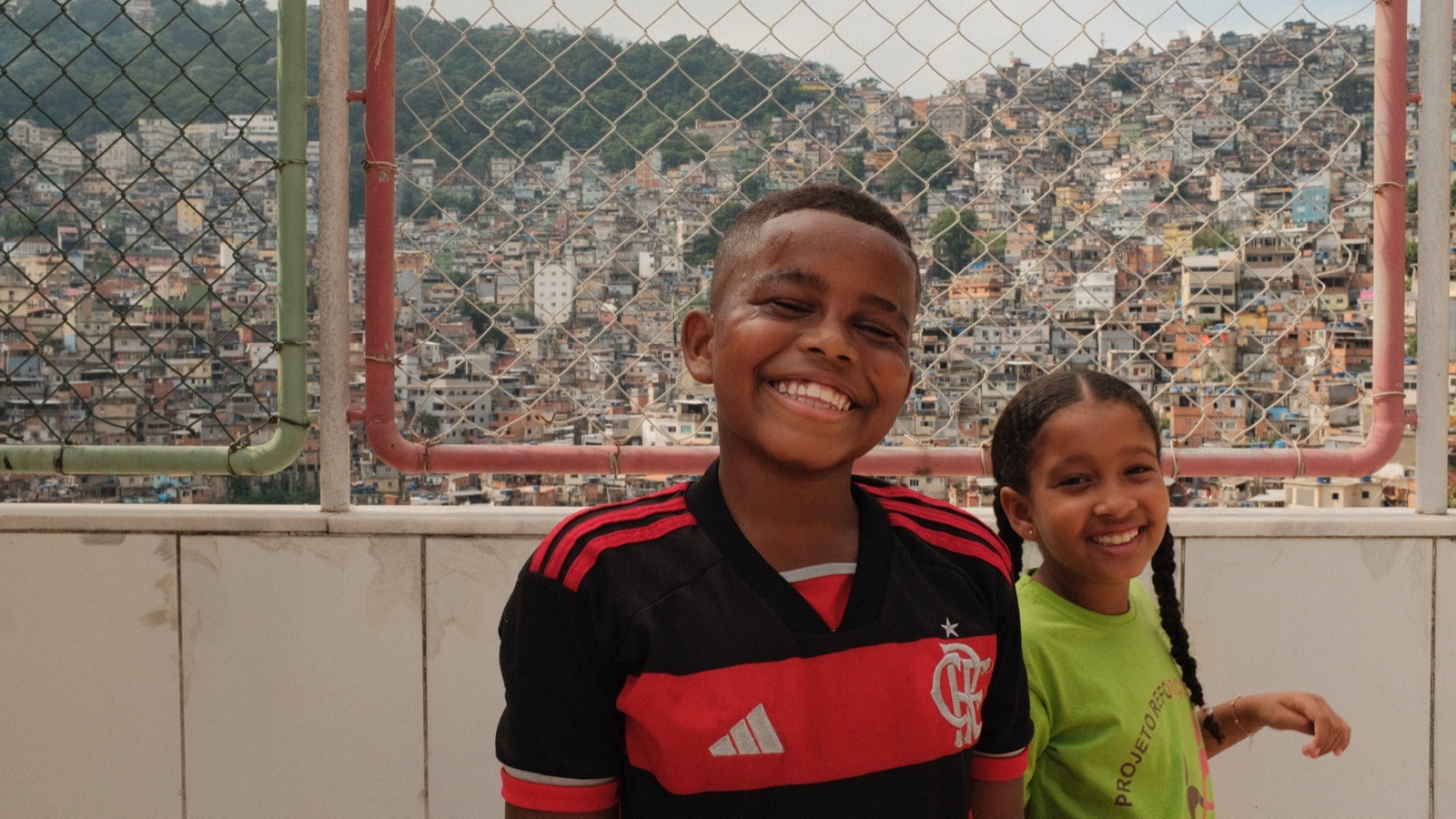 A smiling boy in a Flamengo shirt and a girl in a green T-shirt in front of a chain-link fence, with the houses of the hillside community behind them.