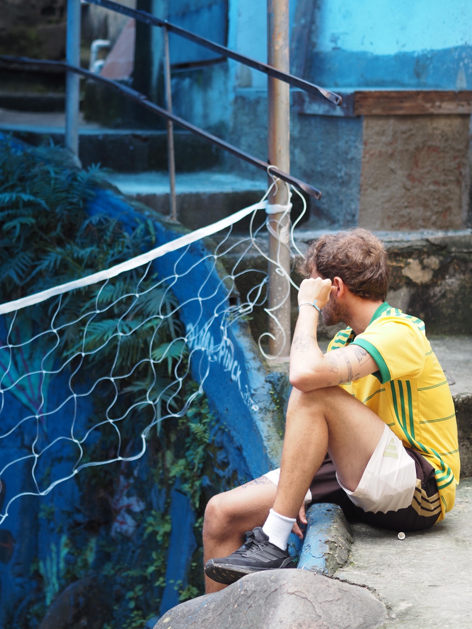 A boy in a yellow shirt sits by a volleyball net in a Vidigal alley.