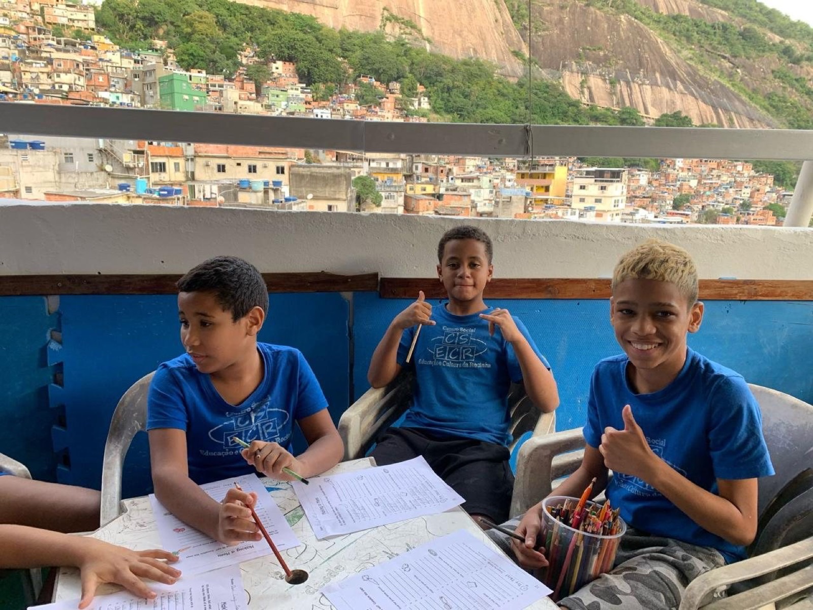 Three boys in blue T-shirts work on English worksheets at a table on a terrace above the hillside community.