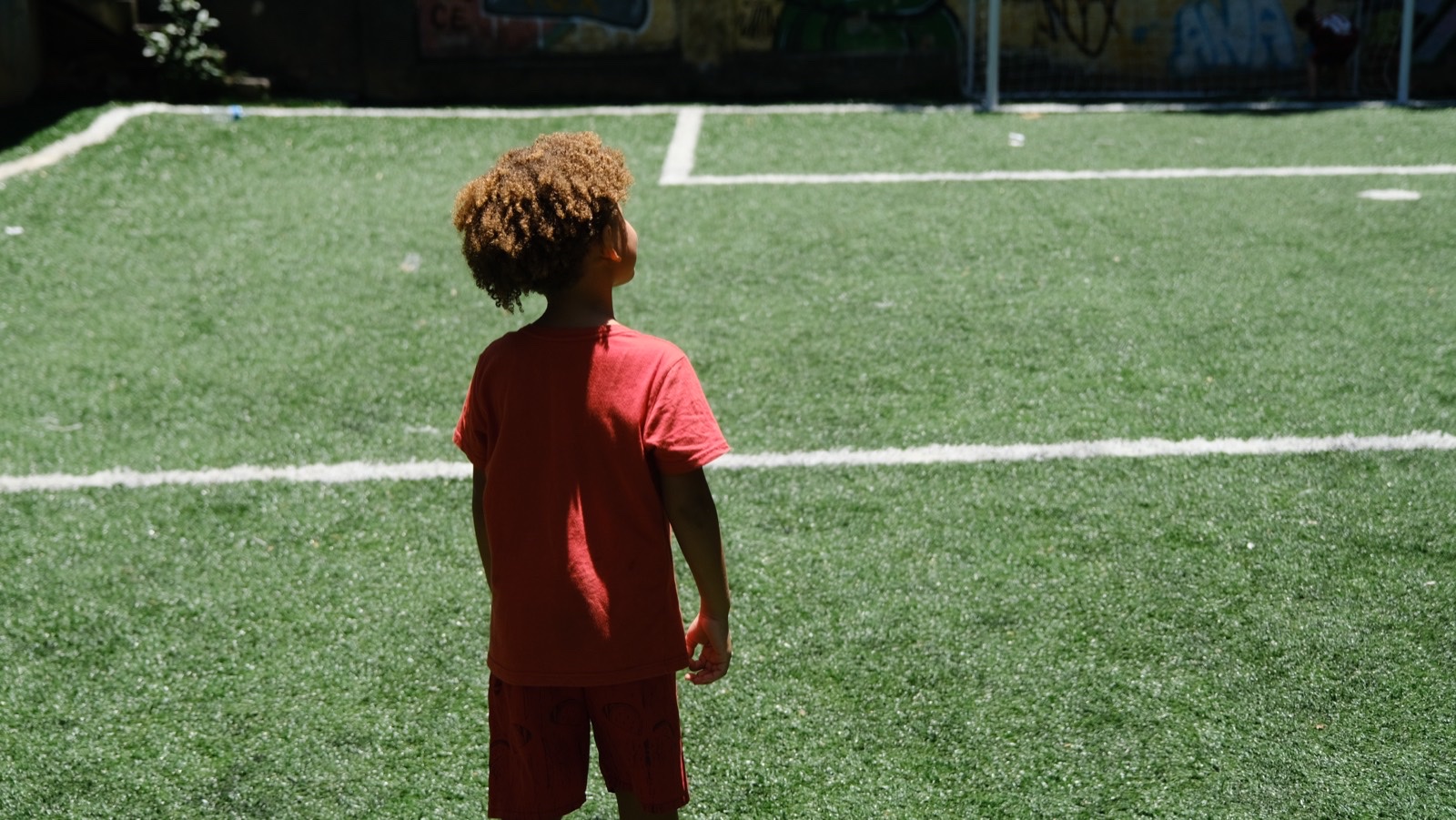 A boy in a red T-shirt standing alone on the artificial pitch, looking out.
