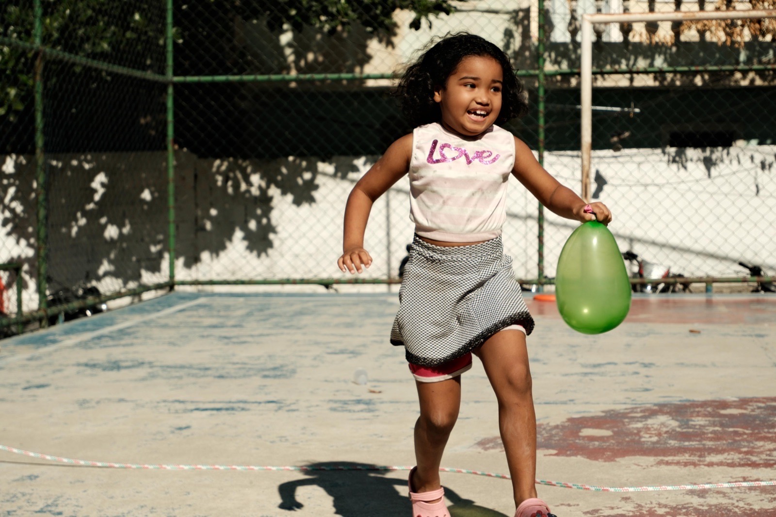 A laughing girl runs across a sports court holding a green water balloon.