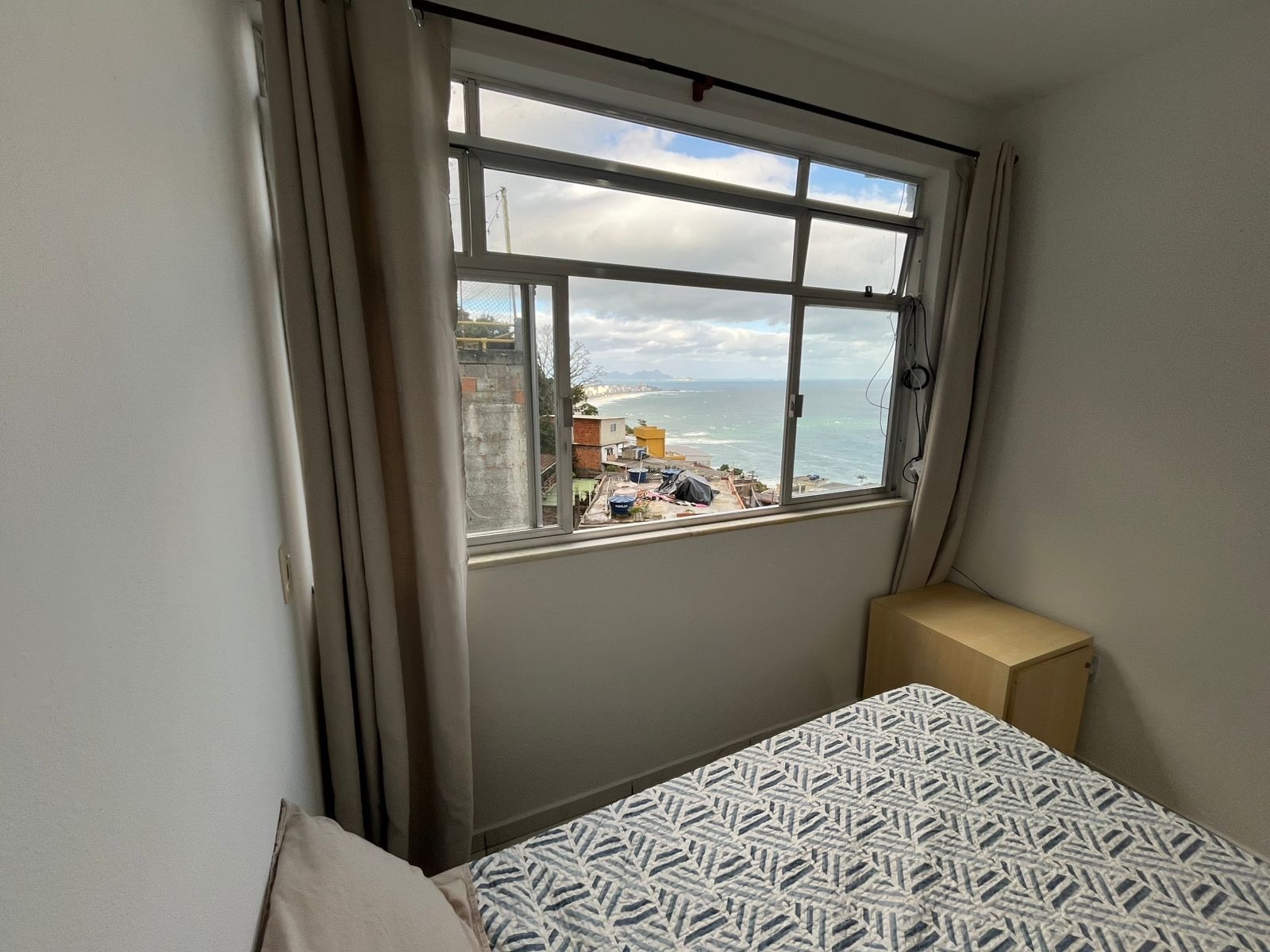 A private room in the co-living space: a double bed beside a large window looking out over the rooftops of Vidigal to the sea.