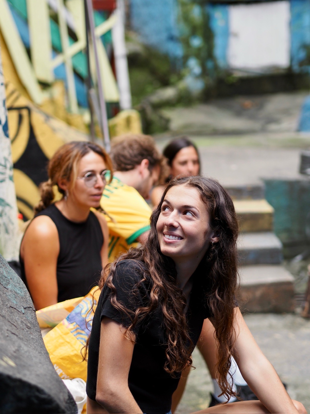 A volunteer with long dark hair smiles up at a friend while sitting with others on painted steps in Vidigal.