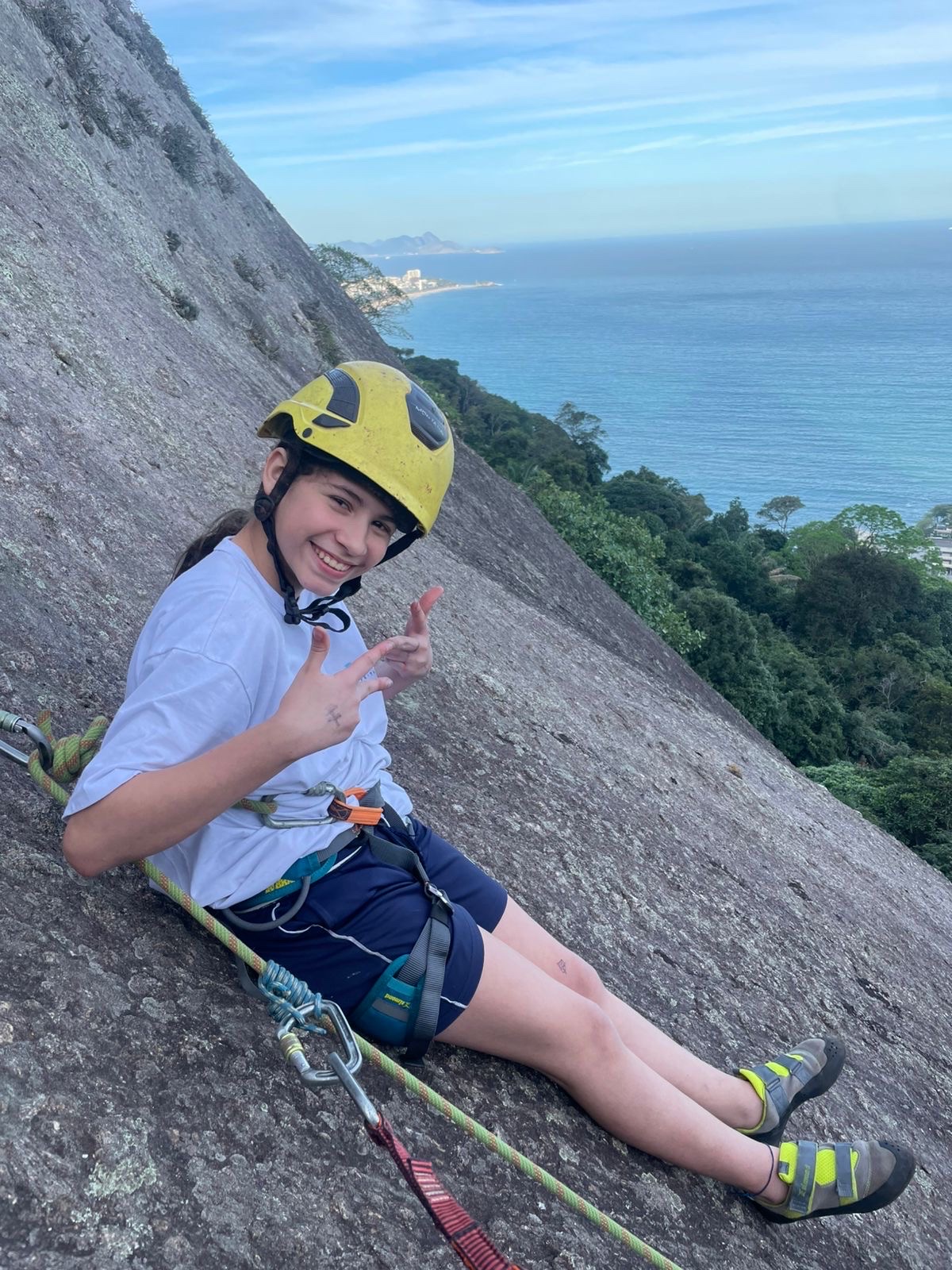 A smiling young climber in a yellow helmet rests on a granite slope high above the sea.
