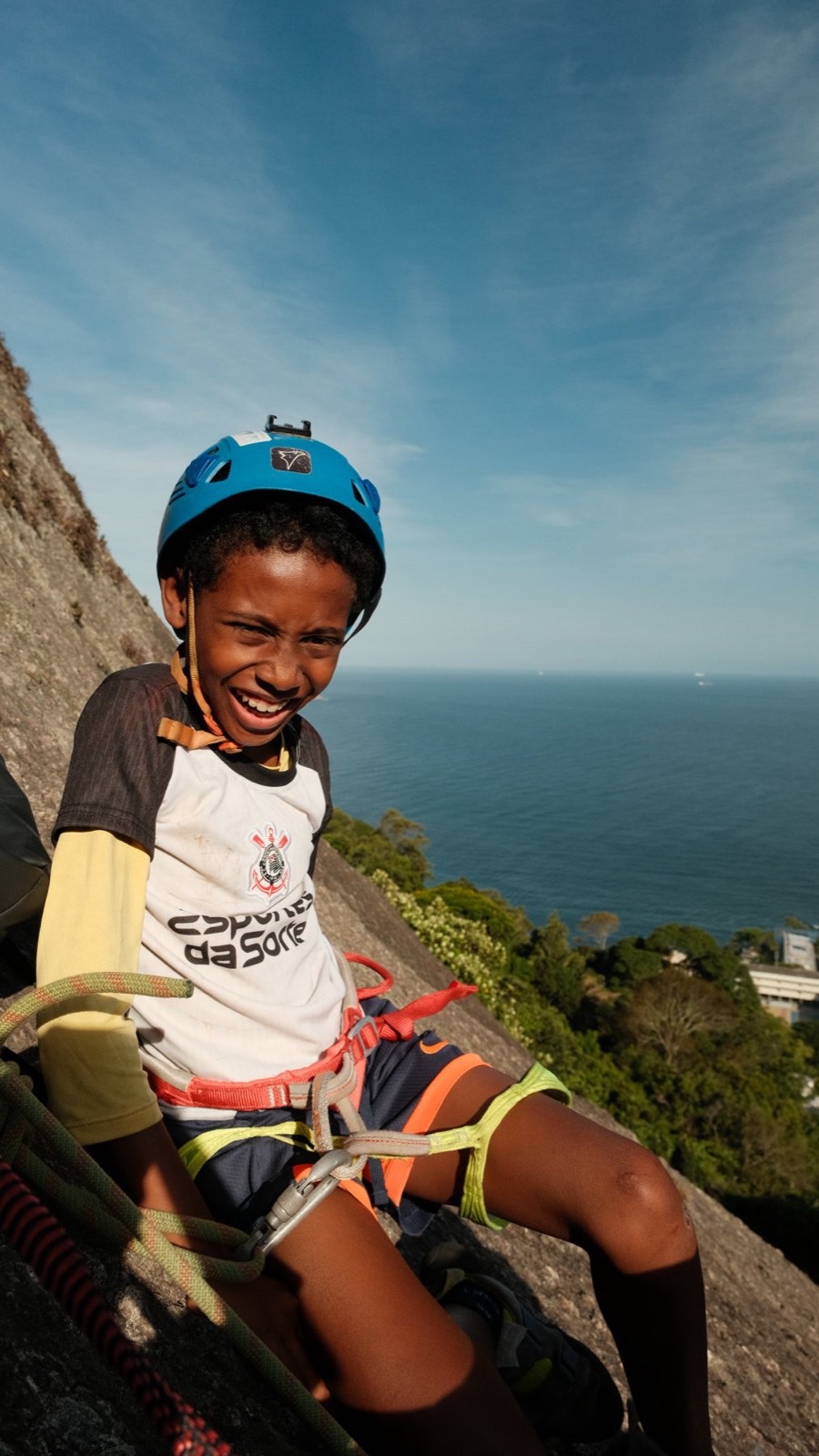 A laughing child in a blue climbing helmet and harness on a rock face high above the sea.